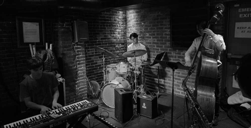 Student jazz trio performing live at Wally’s Cafe Jazz Club, featuring keyboard, drums, and upright bass in an intimate brick-walled setting.